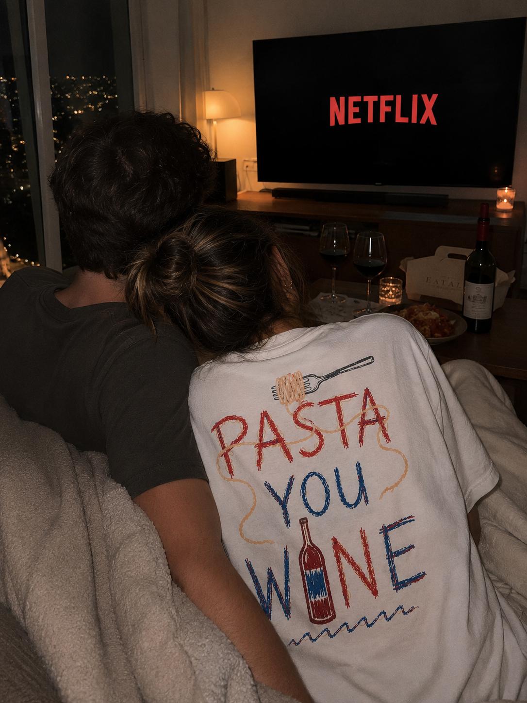 A couple curled up under a blanket watching a film with red wine and leftovers